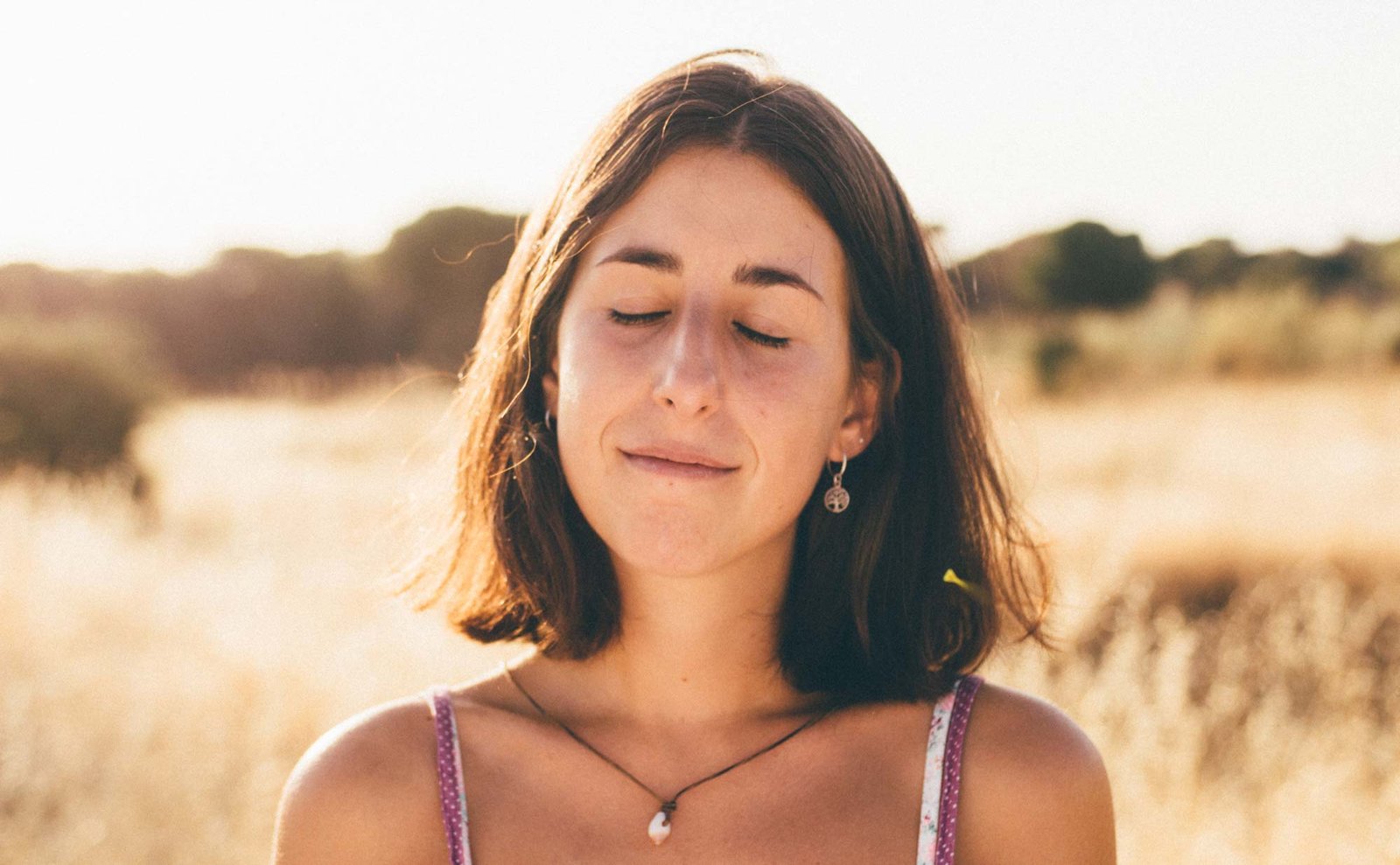 Retrato de una mujer joven de pelo castaño, con los ojos cerrados y una leve sonrisa, a la luz cálida de un campo de hierba seca.
