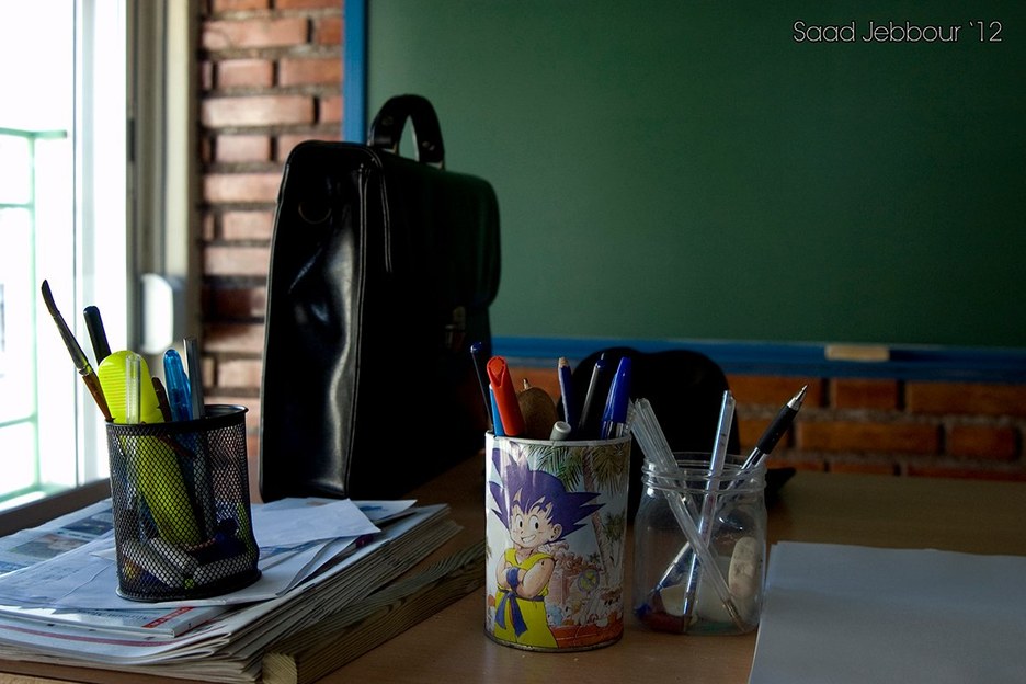 Mesa de profesor con un maletín negro, vasos con bolígrafos (uno con un dibujo de Goku) y periódicos, delante de una pizarra verde y una pared de ladrillo.