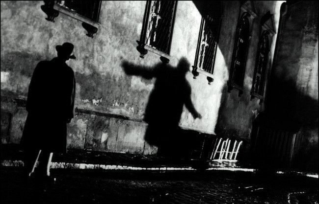 Fotograma en blanco y negro de El tercer hombre: de noche, en una calle empedrada, un hombre con sombrero y gabardina observa la sombra alargada de otra figura proyectada sobre una pared.