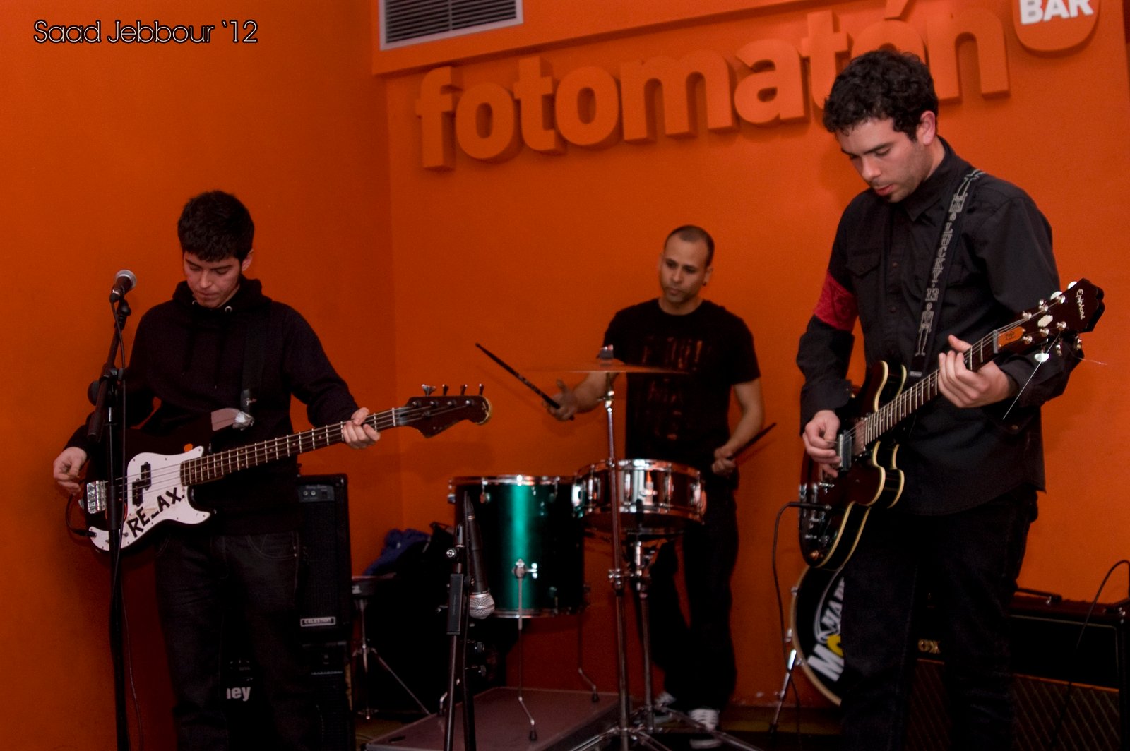 El grupo al completo, bajo, batería y guitarra, tocando ante la pared naranja.
