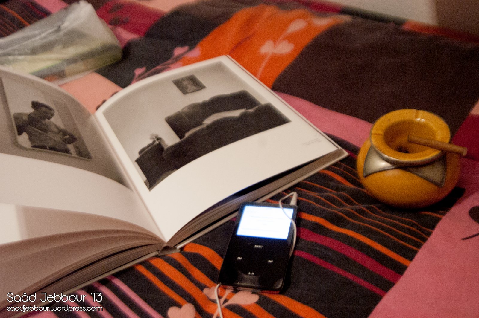 Un libro de fotografía abierto sobre una colcha de rayas, junto a un reproductor de música y un cenicero amarillo con un purito.